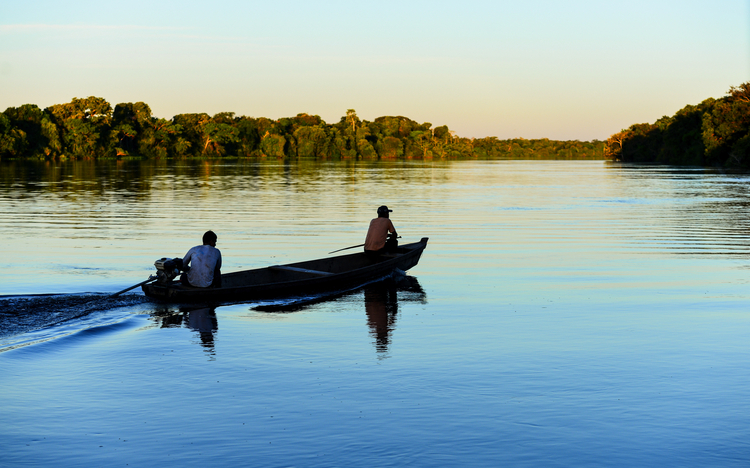 zwei Männer auf einem Boot bei Sonnenuntergang auf dem von Regenwald gesäumten Fluss Guaporé, in der Nähe des abgelegenen Dorfes Mateguá an der Grenze zu Rondônia, Brasilien